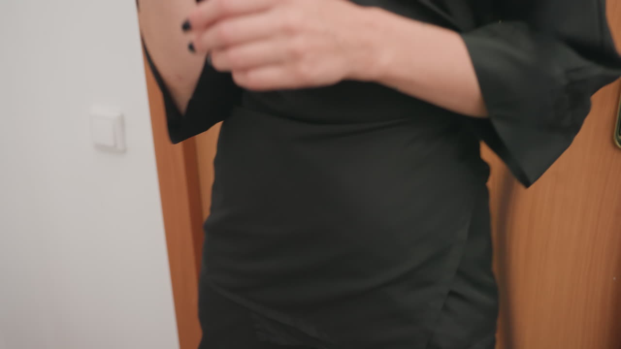 Donna bianca nel backstage che sorseggia acqua vicino alla porta, che posa come una modella, che sistema una veste nera, che controlla i capelli, che indossa sandali vicino alla porta dello studio con espressione rilassata e sicura di sé e un sorriso accennato, spontanea