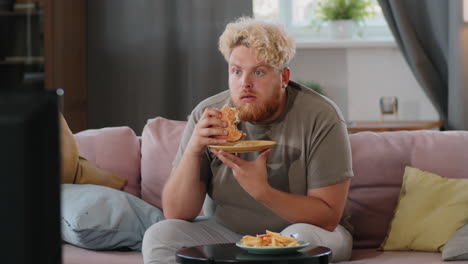 hombre obeso comiendo hamburguesa y amigo mientras ve la televisión en casa