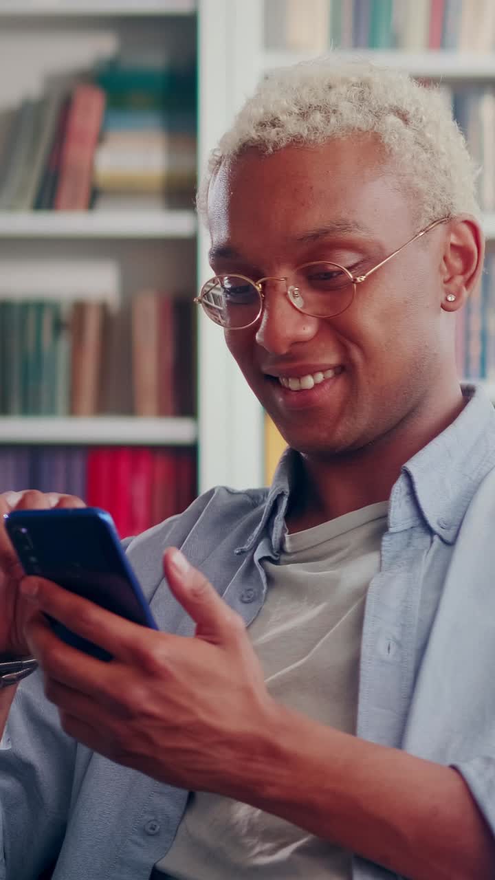 Young smiling african american man in glasses uses smartphone sits in apartment