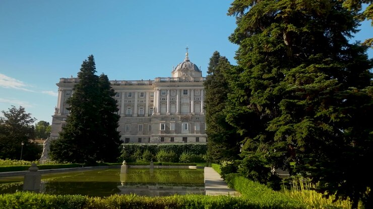 Una vista panoramica del Palazzo Reale di Madrid, Spagna, incorniciato da lussureggianti alberi verdi e una tranquilla piscina riflettente nei Giardini di Sabatini. Ideale per documentari di viaggio e contenuti sul turismo europeo.