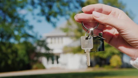 a hand with a keychain in the shape of a small house and keys. against the background of a typical house in the usa suburban style