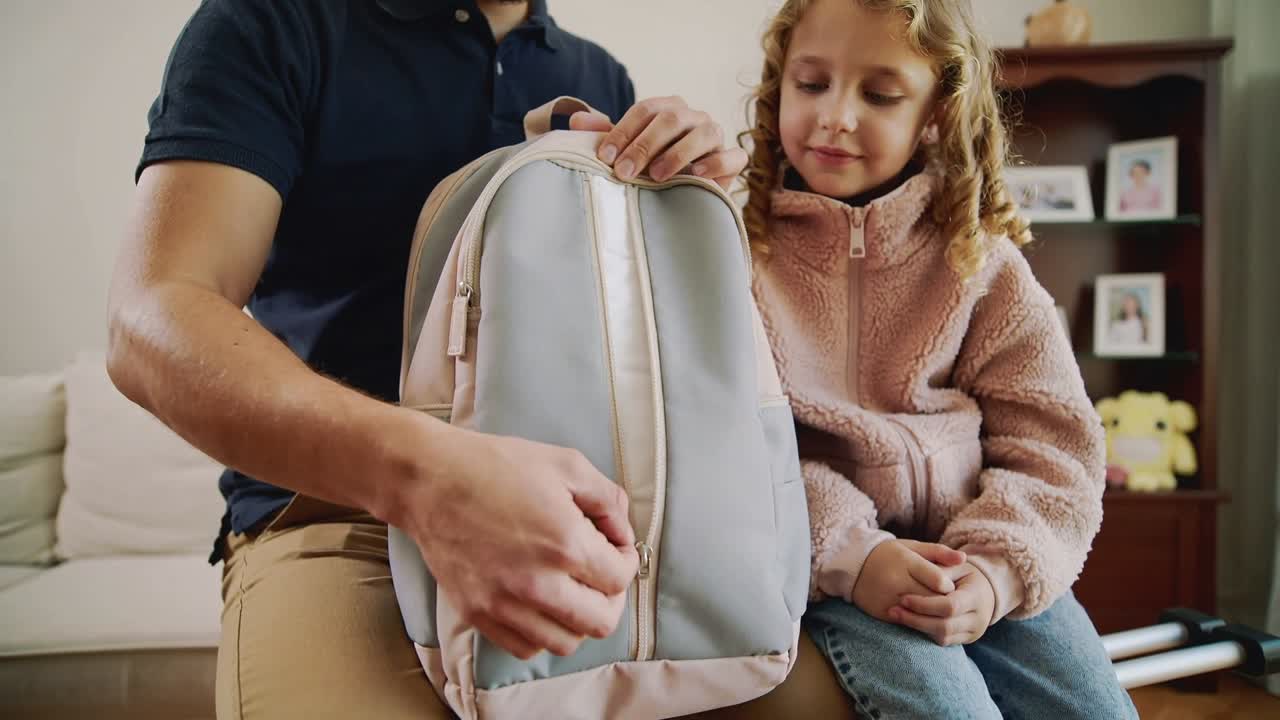 Positioning father in navy polo pulling backpack zipper on couch, girl in pink jacket watching