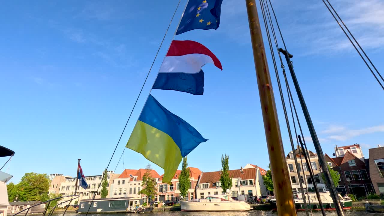 European, Dutch, and Ukrainian flags waving on boat mast, sunny day, canal, historic buildings