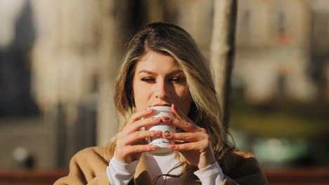 slow motion of a young beautiful blonde woman drinking her coffee from a takeaway coffee cup and smiling, on a sunny morning