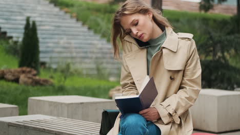 Caucasian female student talking on the phone while writing in a notebook outdoors.
