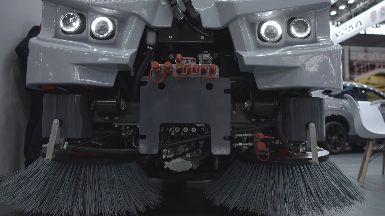 Front-facing industrial street sweeper display inside bright trade show hall, with twin circular lights, broad gray brush assemblies, exposed hydraulic hardware, and heavy central mount surrounded