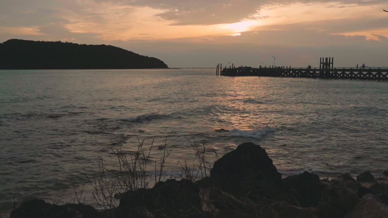 каменистый пляж с пирсом и силуэтами островов во время заката солнца