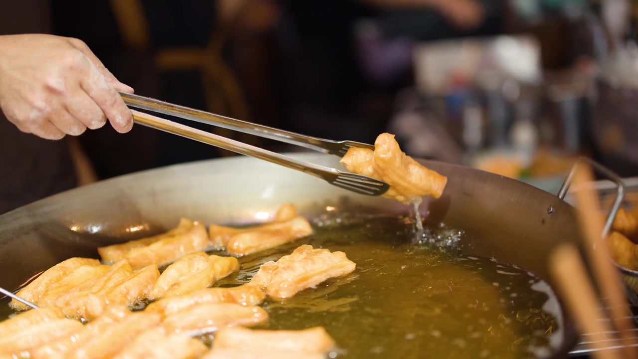Gros plan de mains utilisant des pinces métalliques pour faire frire des bâtonnets de pâte dorés dans de l'huile chaude.