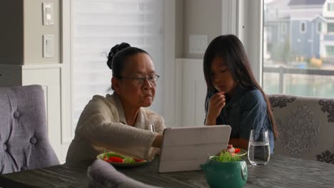 Front-view-of-cute-asian-granddaughter-and-old-grandmother-using-digital-tablet-while-eating-food-4k