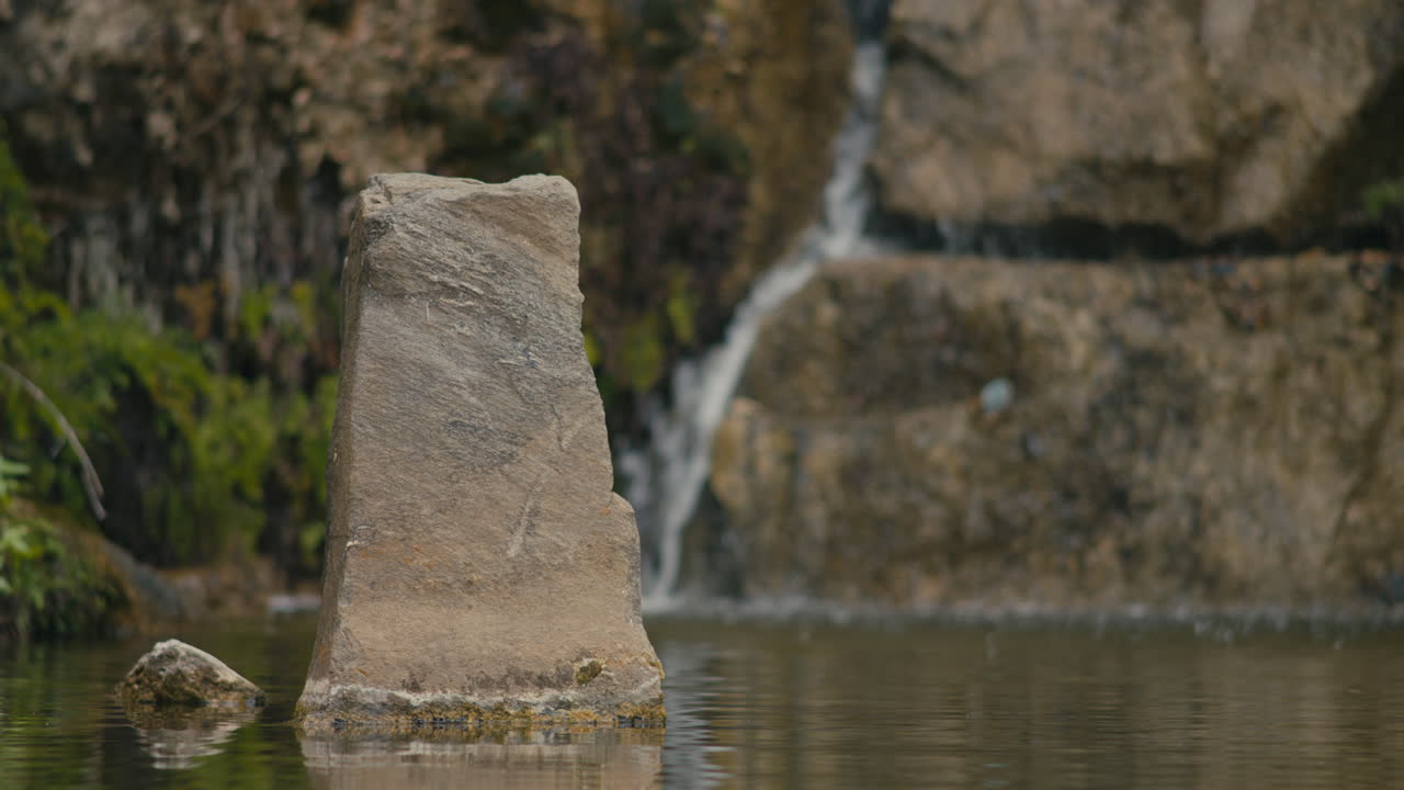 Pilastro di pietra eroso che emerge dall'acqua stagnante di uno stagno, con una dolce cascata che scende sulle rocce sullo sfondo, creando una scena naturale tranquilla.