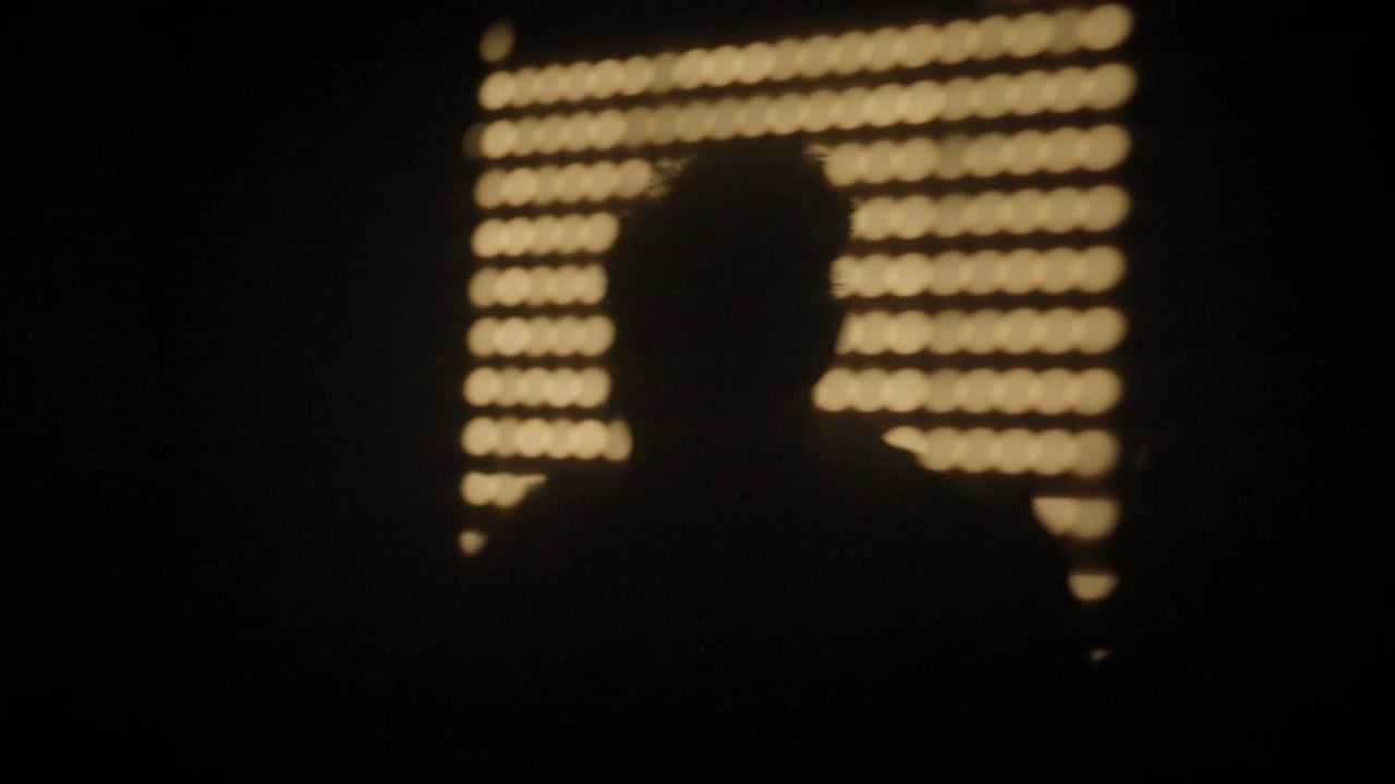 Person's dark silhouette standing in front of a golden bokeh pattern on a black background, creating a mysterious and abstract scene