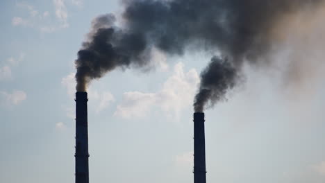 two factory chimney pipes producing heavy dark smoke. industry polluting the atmosphere. cloudy sky backdrop.