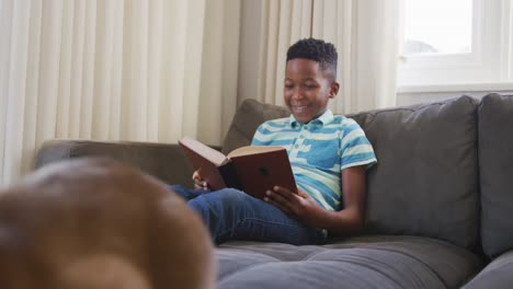 Happy-african-american-boy-sitting-on-couch-reading-book-and-smiling,-pet-cat-sitting-in-foreground