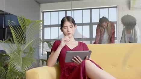 thoughtful asian casual businesswoman using tablet, diverse colleagues in background, slow motion