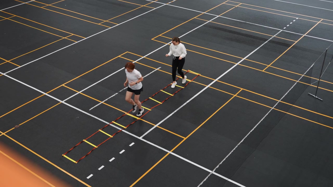 White PE teacher guiding student through badminton ladder drill on indoor court, energetic school gym atmosphere with marked lines and polished floor, emphasis on coordination timing and fun fitness