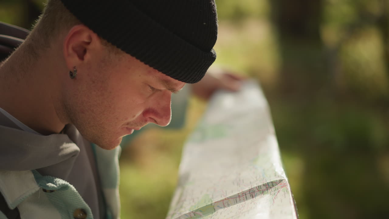 white backpacker unfolding map under trees, furrowed brow scanning creased paper, searching for landmark, soft morning light filtering through leaves, backpack and beanie present, palpable tension