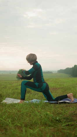 side view of lady in green and black suit outdoors in a vast grassy field practicing low lunge pose on a yoga mat, under cloudy sky with trees in the background and boots with green cloth nearby