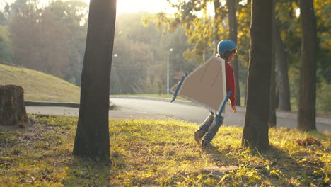 niño pequeño con casco y suéter rojo con alas de avión de cartón corriendo alrededor de los árboles en el parque en un día soleado y jugando como piloto