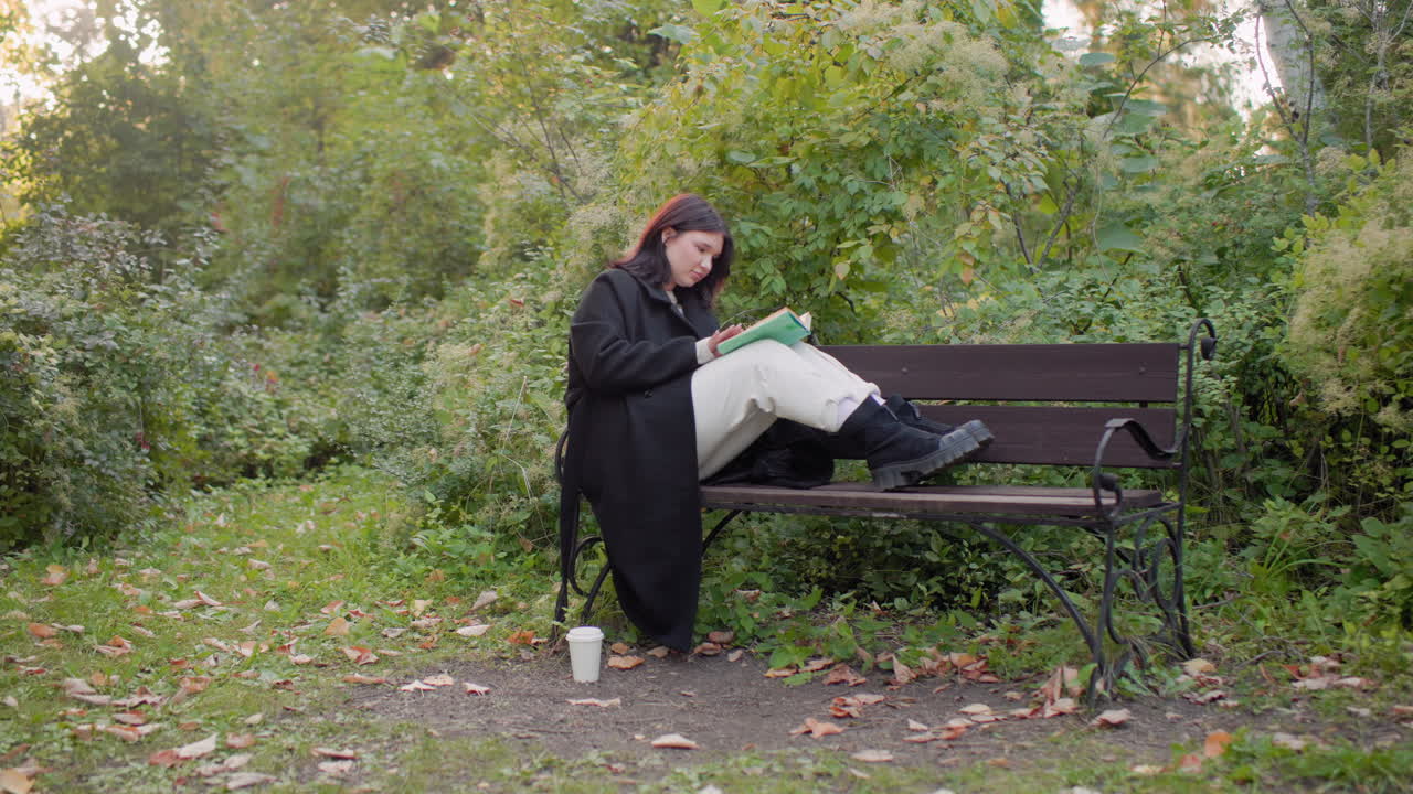 Pensive girl on bench with legs up reading book in green park, sipping coffee from paper cup on ground, cozy black coat and boots, quiet autumn nature, soft light, relaxed mood, slow study moment