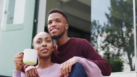 Happy-african-american-couple-embracing-and-relaxing-with-coffee-outside-house