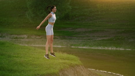 Squats-and-jumps-up.-A-woman-does-exercises-for-the-muscles-of-her-thighs-and-legs-on-the-grass-in-a-Park-near-the-lake.-Slim-beautiful-figure.-Fitness-marathon.-Working-on-your-body