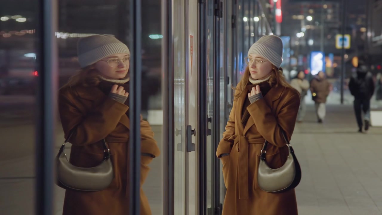 Donna bianca cammina accanto a una facciata di vetro, pendolare solitaria in soprabito marrone che si sistema la sciarpa e porta una borsa, vetrine riflesse e traffico sfocato creano una scena notturna metropolitana ritmica.