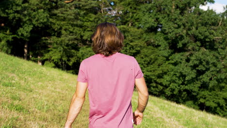 Joven-Con-Camiseta-Roja-Y-Cabello-Largo-Camina-A-Cámara-Lenta-En-Un-Prado-Verde-Hacia-Un-Bosque-En-Un-Día-Soleado-De-Verano