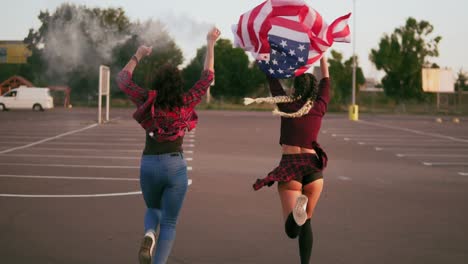 vista de atrás de jóvenes felices chicas hipster estadounidenses corriendo mientras sostienen la bandera estadounidense y soltando granada de bomba de humo con