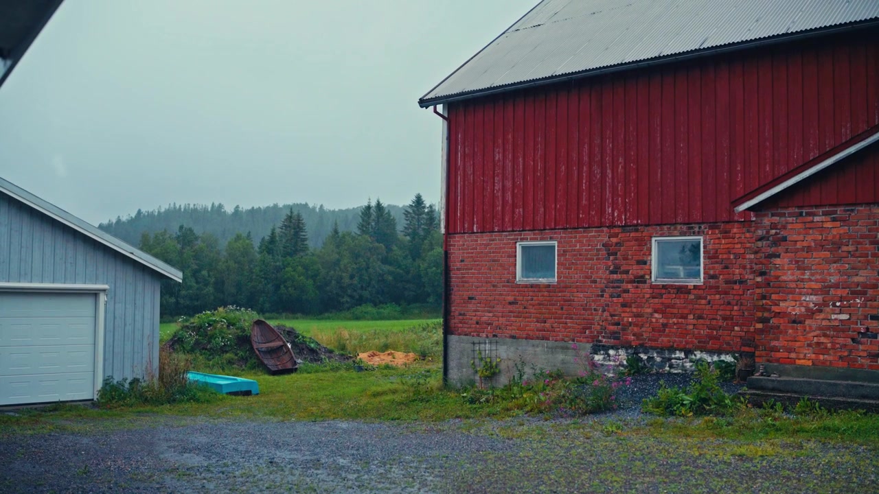 En rød lade og en grå garage står i et regnfuldt landområde omgivet af græs og træer - håndholdt optagelse.