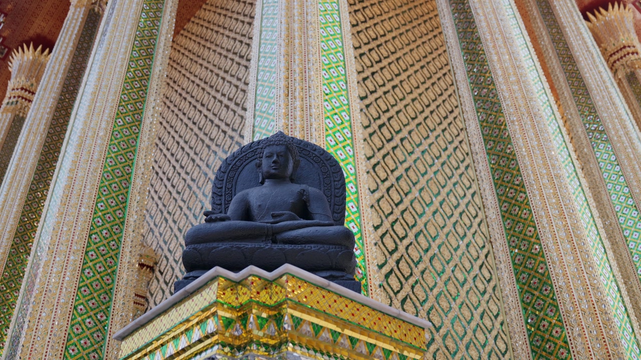A black stone Buddha statue seated in meditation posture is displayed against the ornate and colorful patterned walls of a Thai temple