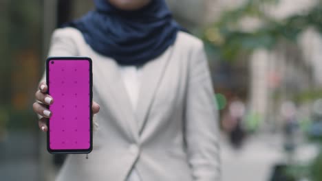 close up of muslim businesswoman holding mobile phone with blank screen to camera standing outside office in city