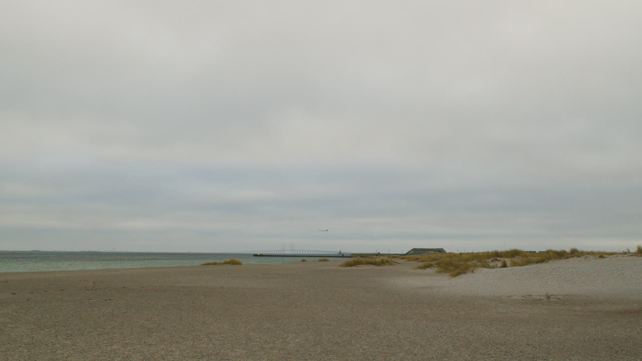 Laaja ja minimalistinen panoraamakuva suuresta, autiosta hiekkarannasta. Pieniä hiekkadyynejä, joissa on harvakseltaan keltaista rantakasvillisuutta, näkyy etualalla ja keskialalla.
