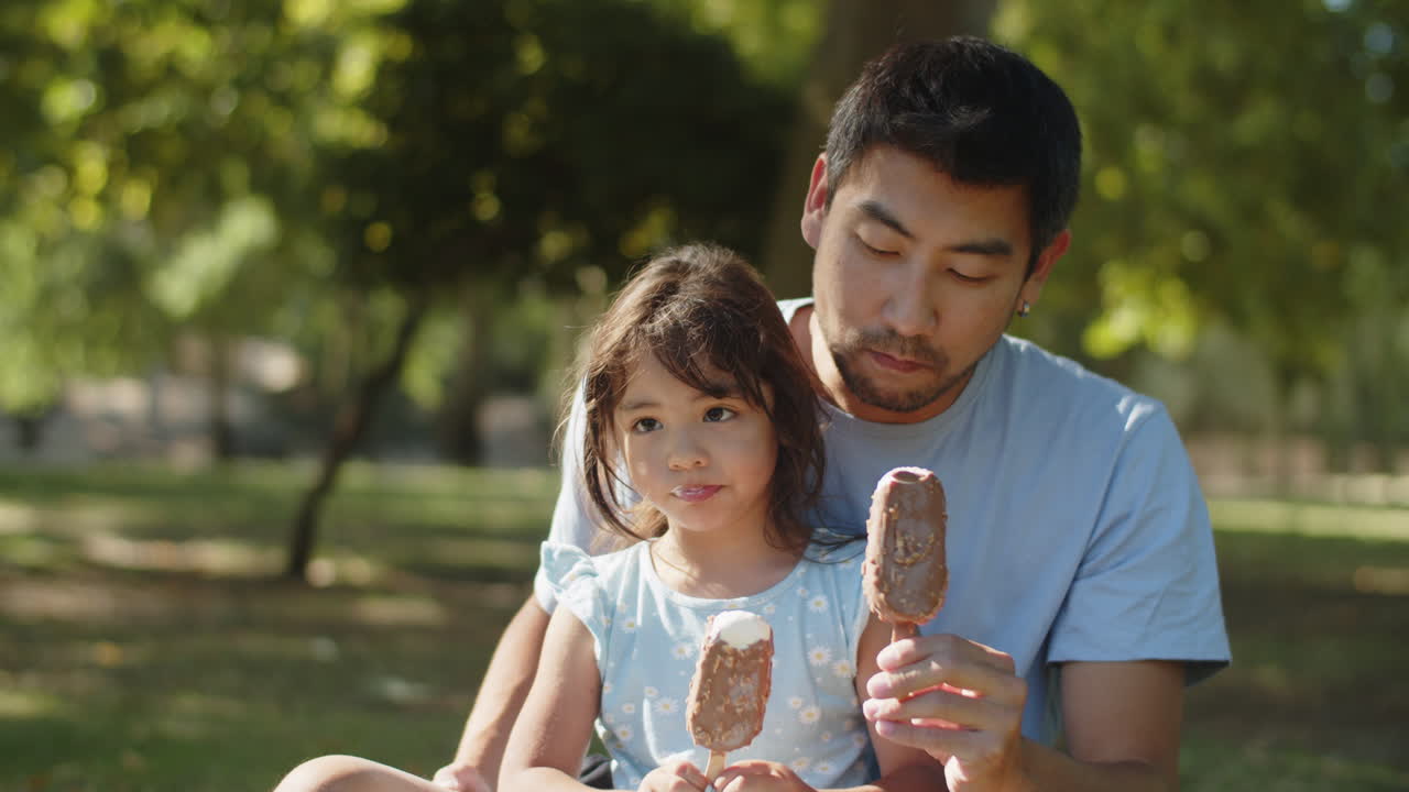 счастливые азиатские отец и дочь едят шоколадное мороженое в парке