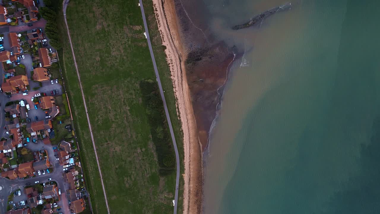 Birds-eye aerial drone moves forward over Weston Shore Southampton, revealing coastal pathway, autumn trees, and shoreline bathed in golden sunset light with calm scenic atmosphere