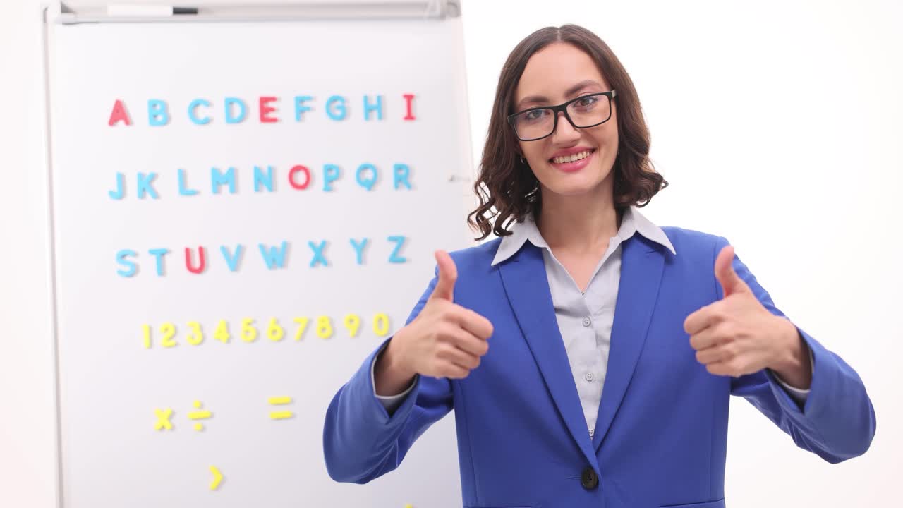 Teacher giving thumbs up in front of whiteboard with alphabet and numbers