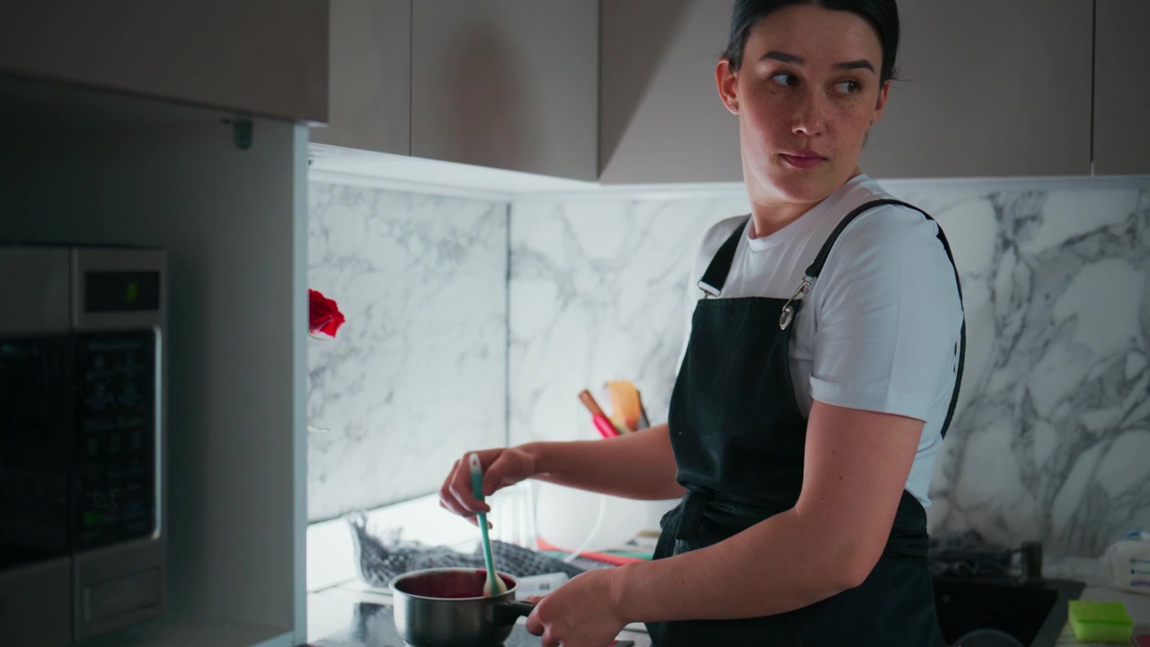 Una donna con un grembiule mescola una pentola in una cucina moderna. Si concentra intensamente sulla sua cucina, la sua espressione è calma e concentrata. Un paraschizzi in marmo ed elettrodomestici da cucina la circondano.