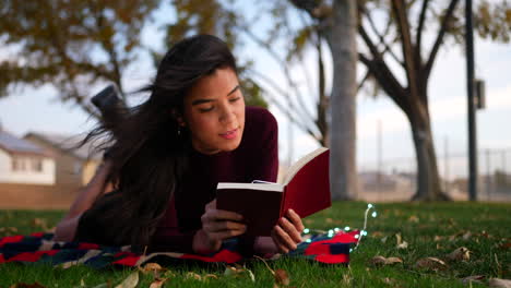 a beautiful young hispanic woman reading a story book or novel in the park with autumn leaves blowing in the wind slide right