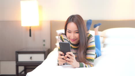 young beautiful asian woman laying on bed, reading messages, articles on mobile phone