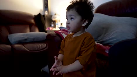 adorable 18-month-old baby boy watches tv in the evening, captivated by the screen and clapping his hands in approval with a curious expression, showcasing the concept of mimicking behavior