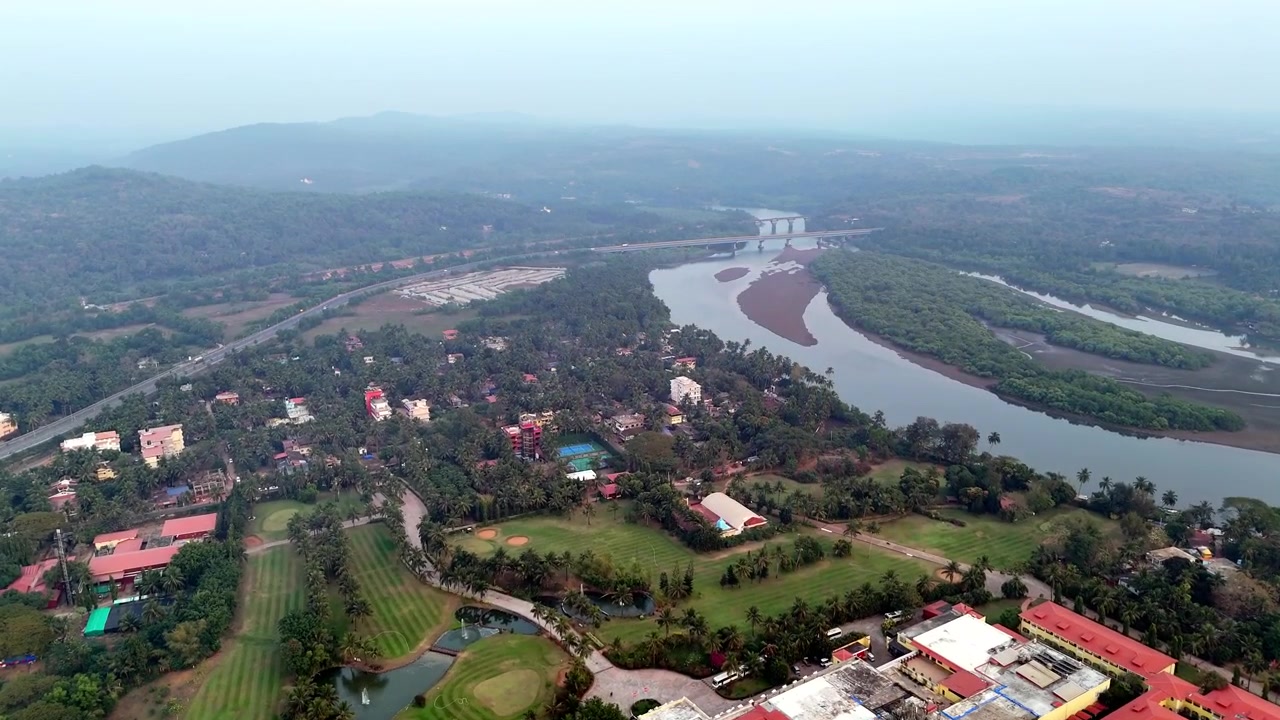 Filmový letecký pohled na Canaconu, ukazující budovy luxusních resortů s červenými střechami, rozsáhlou zeleň, klikatící se řeky a vzdálené kopce splývající s měkkým, mlhavým horizontem. Řeka Talpona a most.