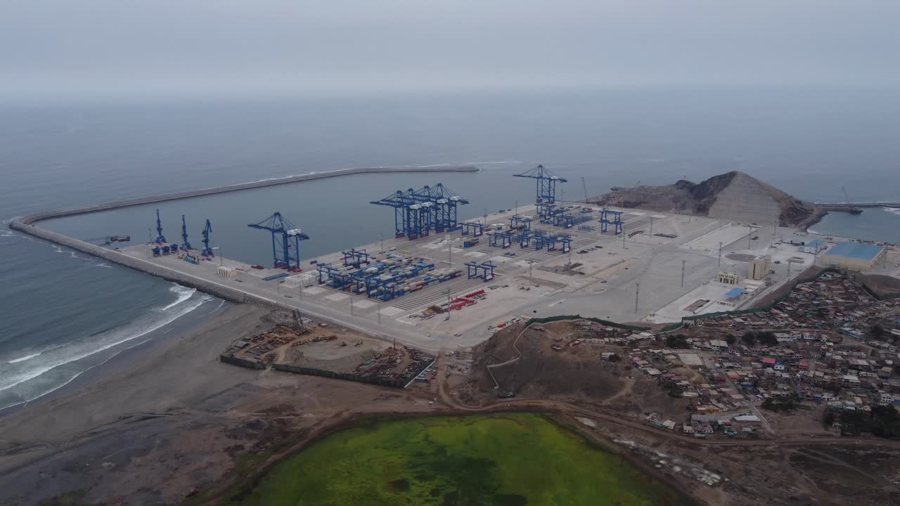 Aerial shot of the construction site of a megaport being built in Peru in the city of Chancay. Drone flies high above while focusing down on the site below with many tall blue cranes in the harbor.