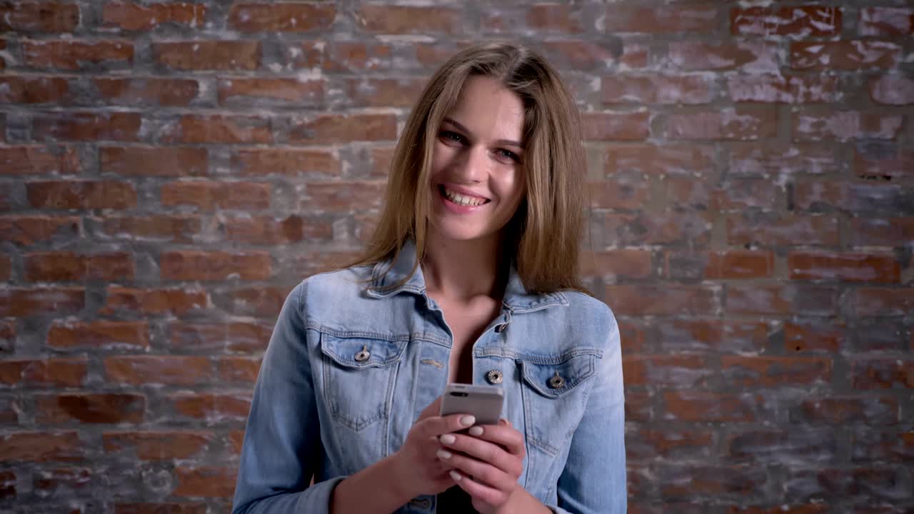 una joven linda está escribiendo un mensaje en su teléfono inteligente, mirando a la cámara, el telón de fondo de ladrillo, el concepto de comunicación