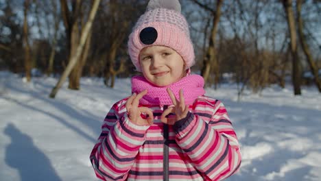 Child-girl-smiling,-showing-ok-sign,-thumbs-up-gesture-on-snowy-road-in-winter-sunny-park-forest