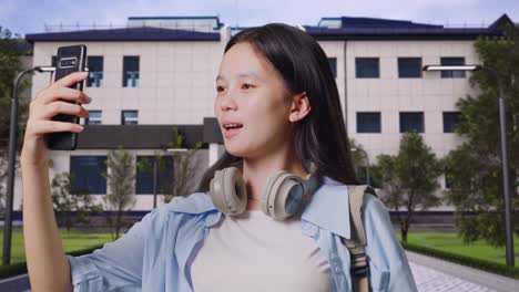 close up of asian teen girl student with a backpack having a video call on smartphone while standing in front of a school building