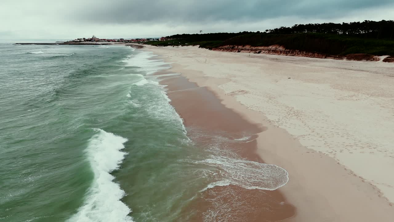 Ondas suaves alcançam uma praia ampla e vazia perto de uma floresta e uma cidade distante.