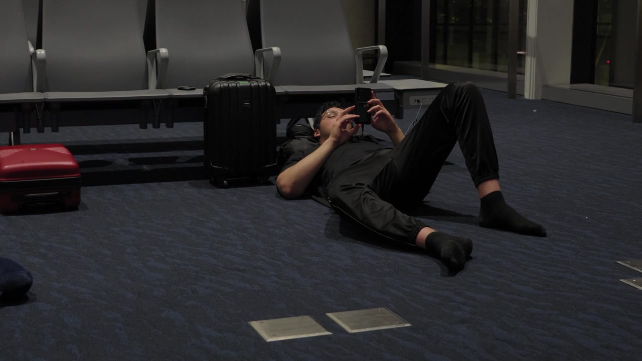 Tired traveler lying on the floor beside luggage using a phone during an overnight layover at an airport terminal