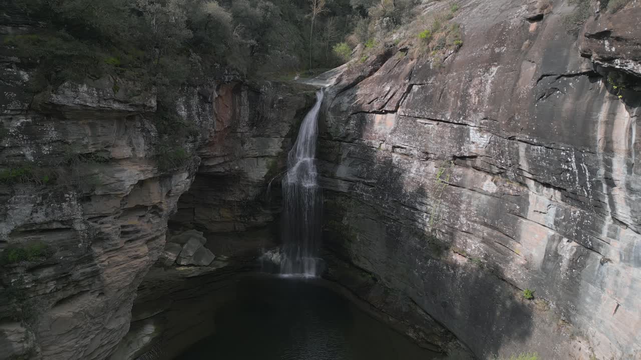 Sunlight illuminates the rugged cliffs surrounding la foradada waterfall as it cascades into a tranquil pool, creating a picturesque scene in the heart of les guilleries, near barcelona