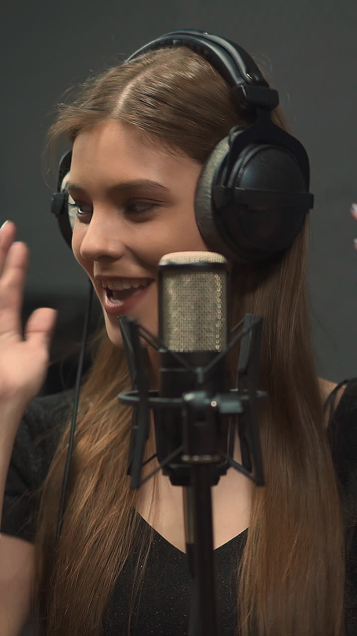 Happy long haired young lady performer with large headphones records new song with contemporary microphone dancing in audio studio close view