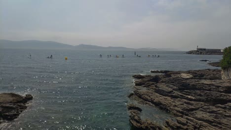 Menschen-In-Einer-Gruppe,-Die-An-Einem-Sonnigen-Tag-Im-Morgengrauen-An-Der-Küste-Mit-Dem-Ruhigen-Meer-Und-Der-Stadt-Im-Hintergrund-Paddle-Surf-Unterricht-Mit-Brötchen-Nehmen