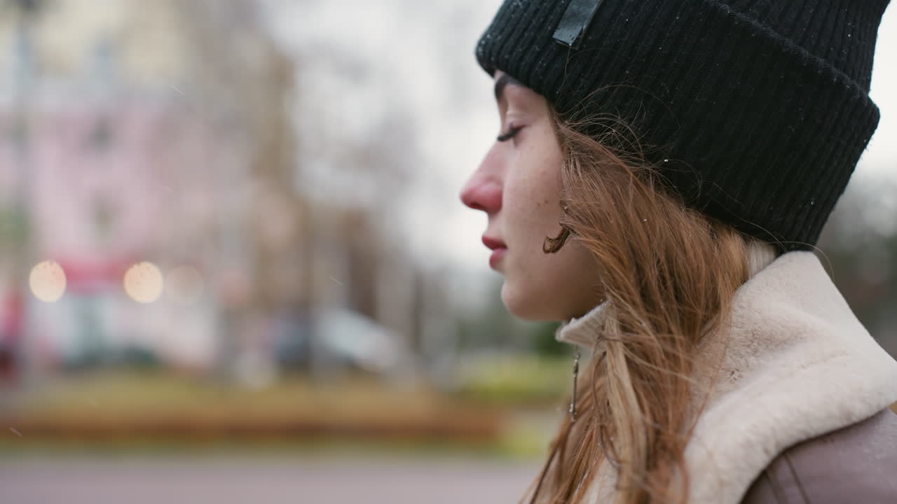 Sideskud af en glad pige med en sort strikhue og brun shearling-jakke, der går udenfor på en kold dag. En blød baggrund med slørede bylys og dæmpede farver skaber en rolig og hyggelig vinterstemning i gaden.